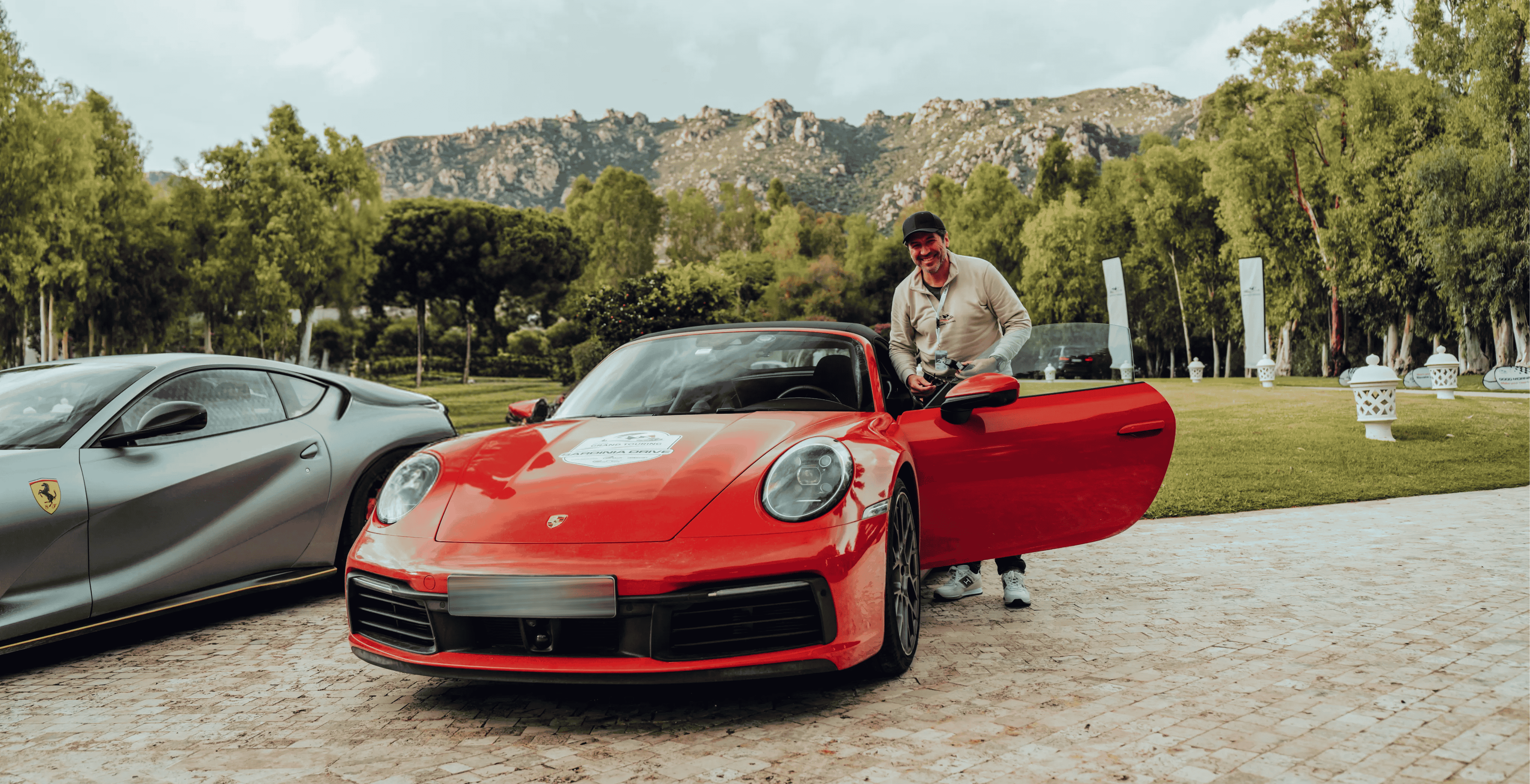 Ein Mann lehnt lachend an einem roten Porsche mit geöffneter Tür, daneben steht ein grauer Ferrari, im Hintergrund Bäume und Berge.