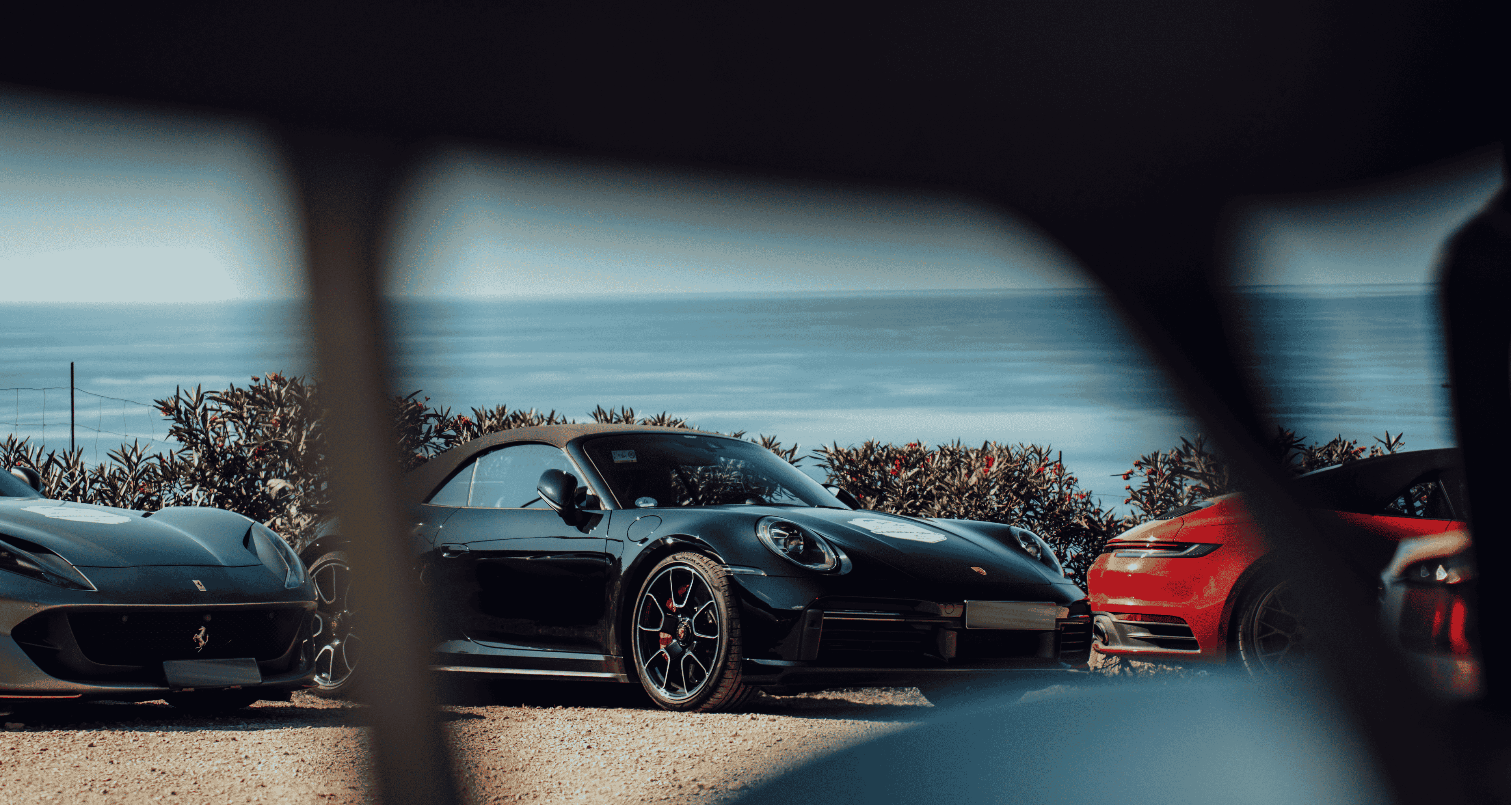 Drei Sportwagen parken draußen auf einem Kiesplatz mit Blick auf das Meer, teilweise durch den Rahmen einer Sonnenbrille gesehen.
