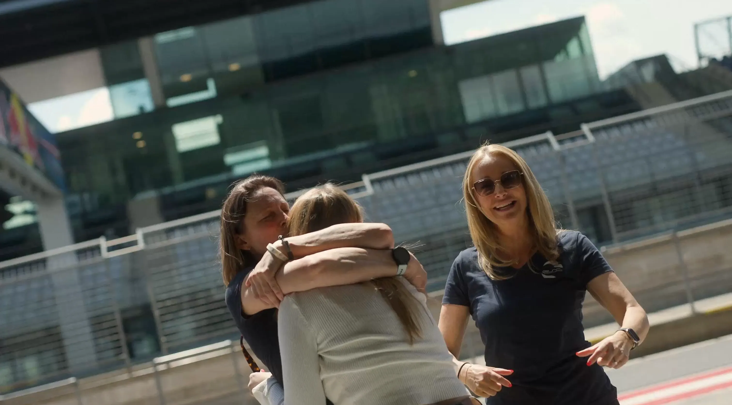 Drei Frauen draußen, zwei umarmen sich, die dritte lächelt in Sonnenbrille.