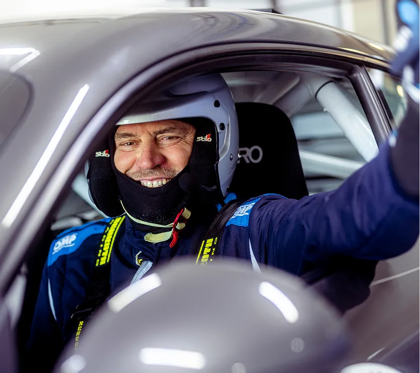 Ein lächelnder Rennfahrer mit Helm und Sicherheitsgurten sitzt im Cockpit eines Rennwagens.