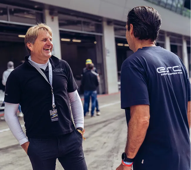Zwei Männer stehen in einer Rennstrecken-Pitlane und unterhalten sich, während im Hintergrund weitere Personen und Garagen zu sehen sind.
