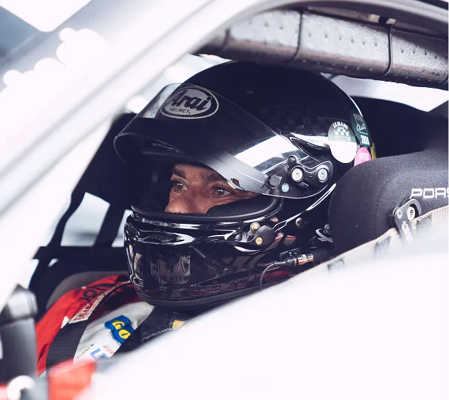 Rennfahrer mit schwarzem Helm sitzt angeschnallt in einem Porsche-Rennwagen.