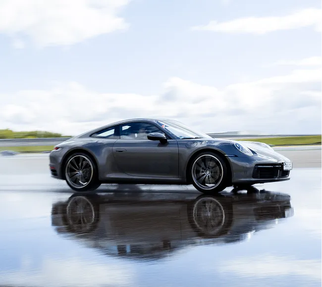 Grauer Sportwagen fährt auf nasser Straße mit Spiegelung im Wasser.