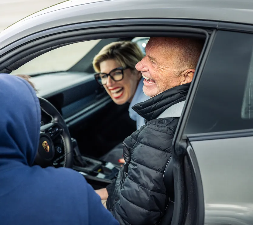 Ein älterer Mann und eine Frau lachen, während sie auf den Rücksitz eines Autos schauen, dessen Tür geöffnet ist.