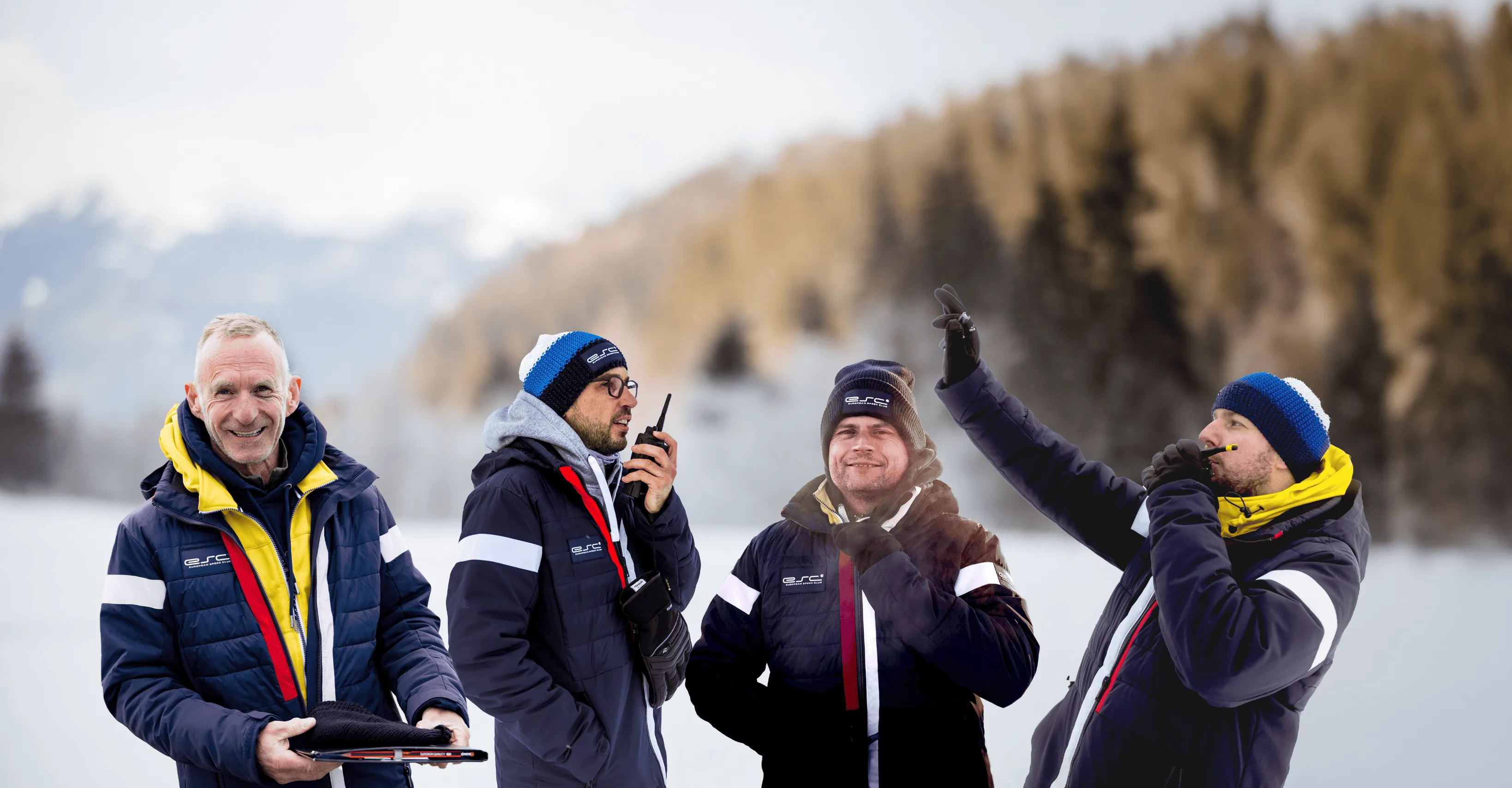 Four men in winter jackets and caps communicate with walkie-talkies over a snow-covered mountain panorama.