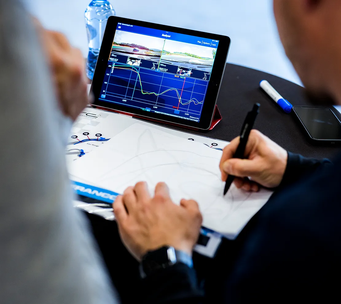 Person draws on a piece of paper next to a tablet, which shows an analysis of racetracks and vehicle data.