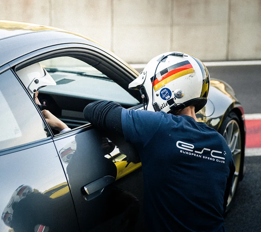 Two people wearing helmets, one sitting in a black sports car, the other leaning towards the window and talking to her.