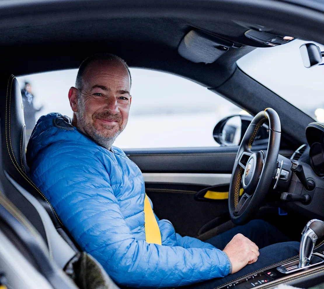 A smiling man in a blue jacket is sitting in the driver's seat of a car with a yellow seatbelt.