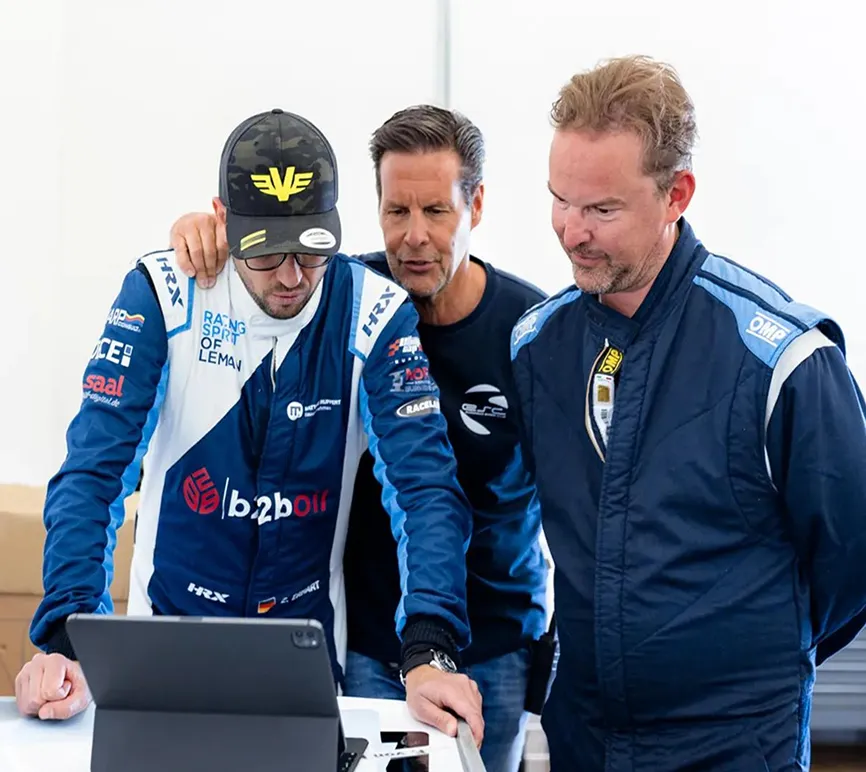 Three men, two in racing suits and one in casual clothes, are looking at a tablet on a table together.