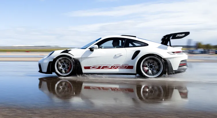 Weißer Porsche GT3 RS fährt schnell über nasse Strecke und spiegelt sich im Wasser.