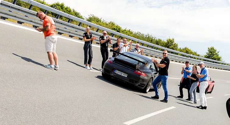 Eine Gruppe von Menschen applaudiert neben einem schwarzen Sportwagen auf einer geneigten Straße mit Metallgeländer.