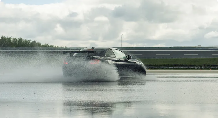 Ein schwarzes Auto fährt auf nasser Fahrbahn und spritzt Wasser hoch, unter einem bewölkten Himmel.