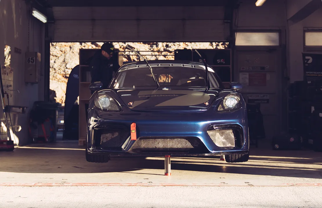 Blauer Porsche-Rennwagen steht in einer Garage auf Stützen mit geöffneter Motorhaube und einem Mann im Hintergrund.
