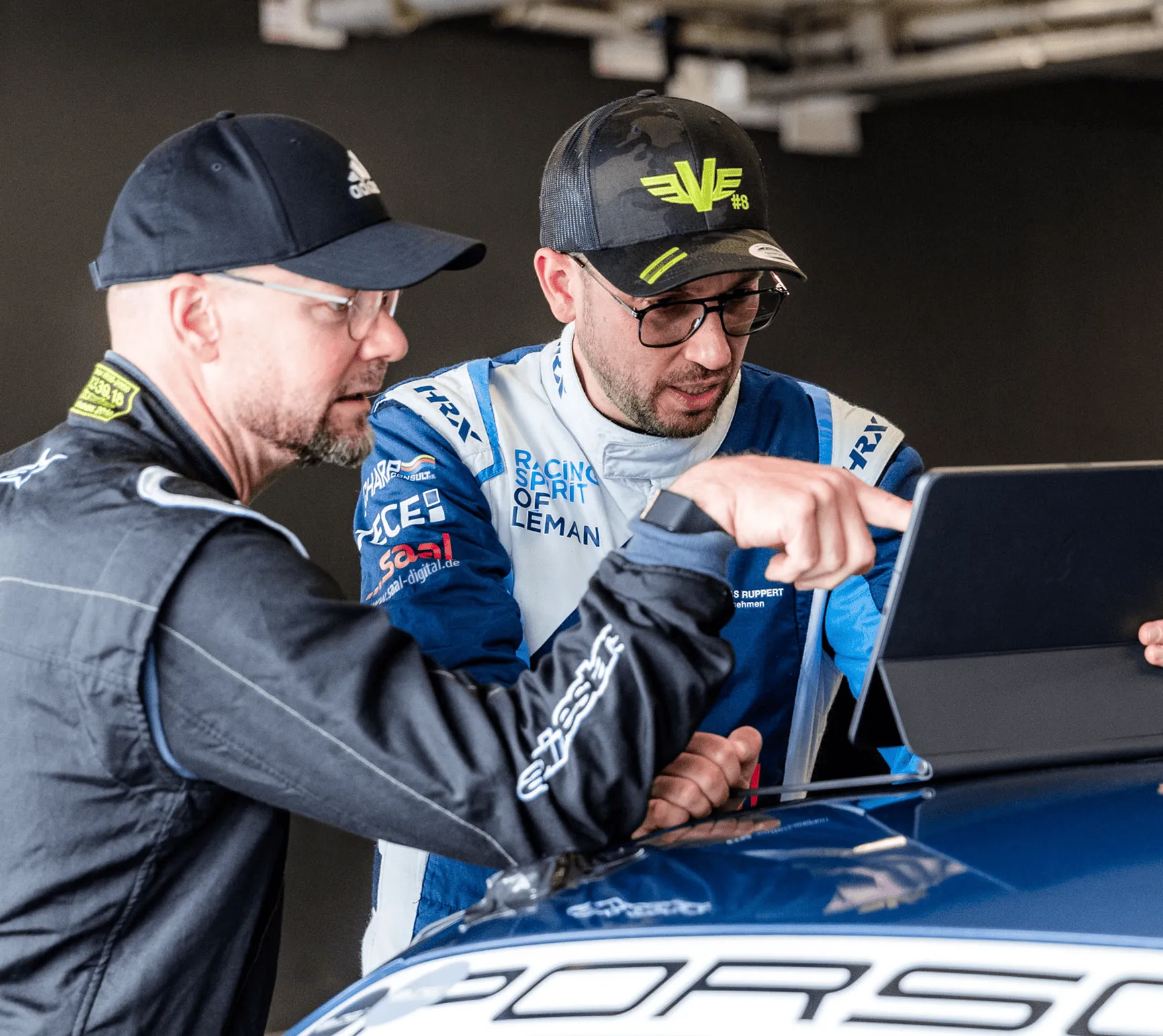 Two men wearing motorsport clothes, one pointing at a tablet, next to a blue Porsche car.