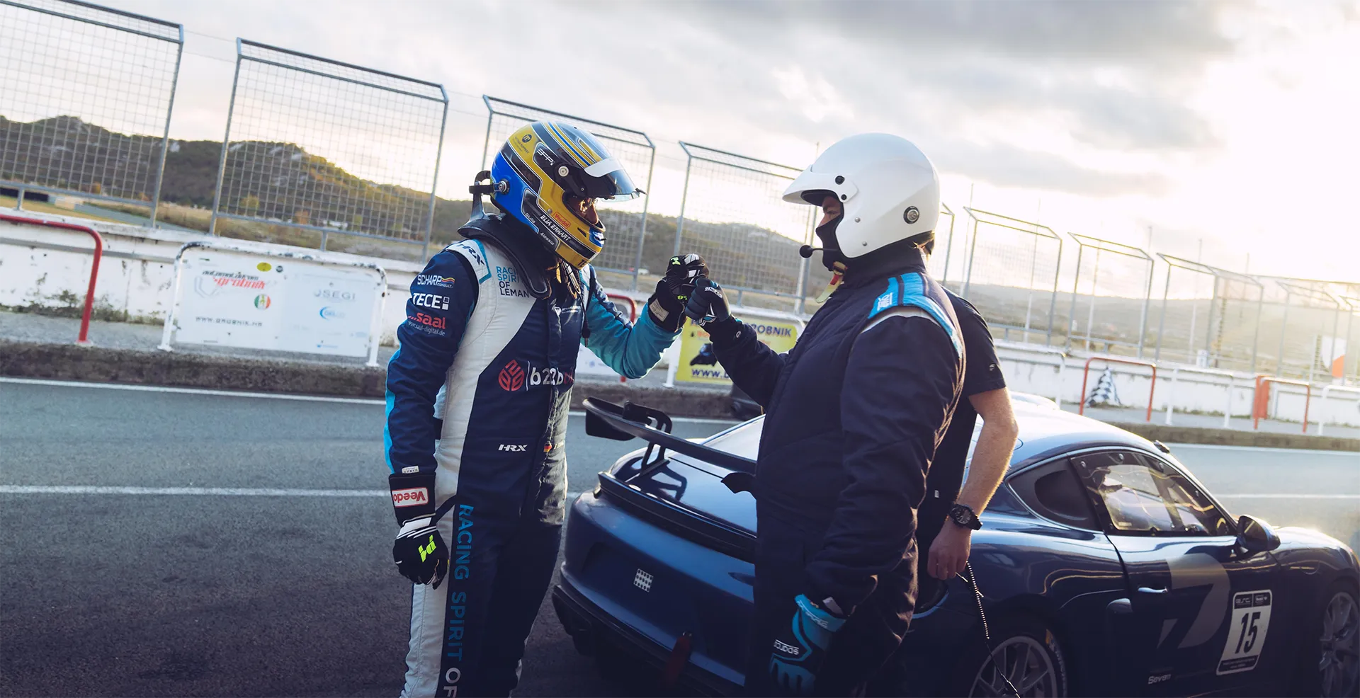 Zwei Rennfahrer in Schutzhelmen und Anzügen geben sich auf einer Rennstrecke am Heck eines blauen Sportwagens einen Faustgruß.