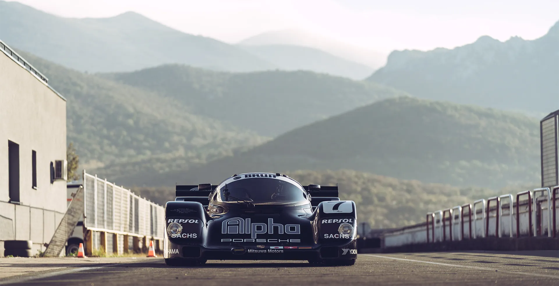 Schwarzes Alpha Porsche Rennfahrzeug auf einer Rennstrecke mit Bergen im Hintergrund.