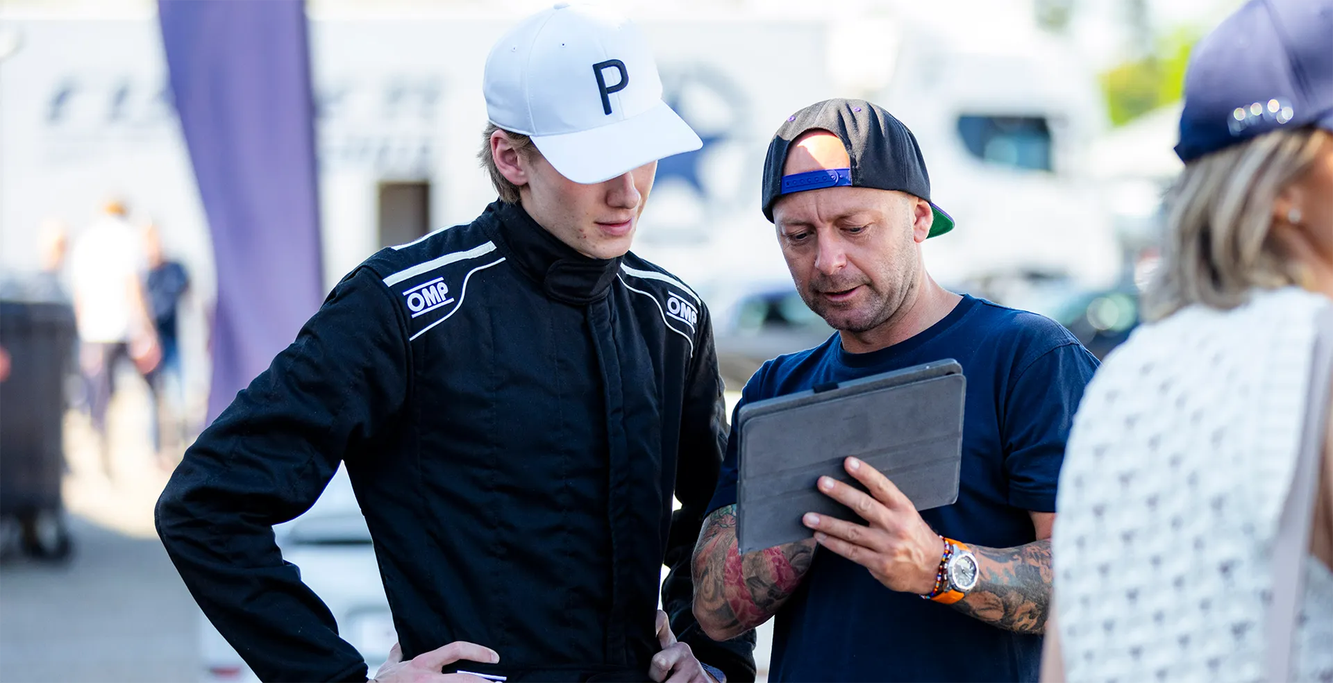 Zwei Männer stehen draußen, einer trägt einen Rennanzug und eine weiße Mütze, der andere zeigt etwas auf einem Tablet.