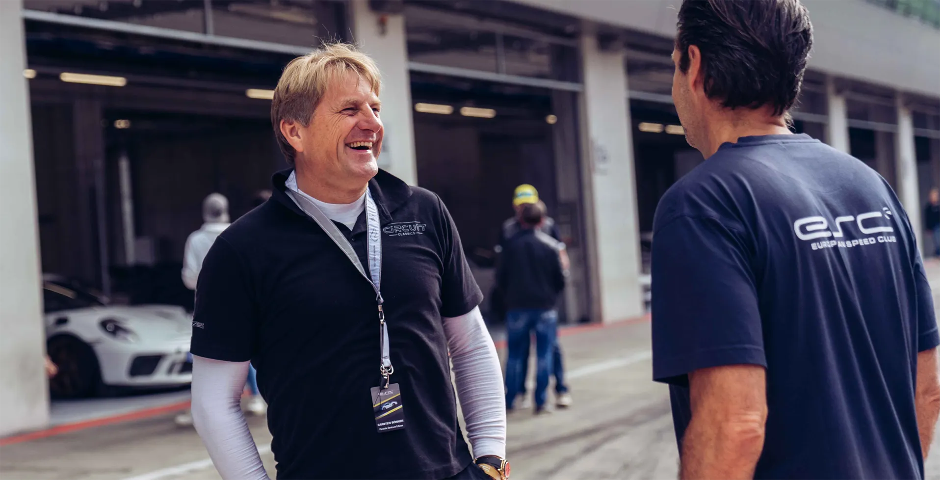 Zwei Männer unterhalten sich lachend außerhalb einer Rennstrecken-Garage, ein weißes Sportauto ist im Hintergrund zu sehen.
