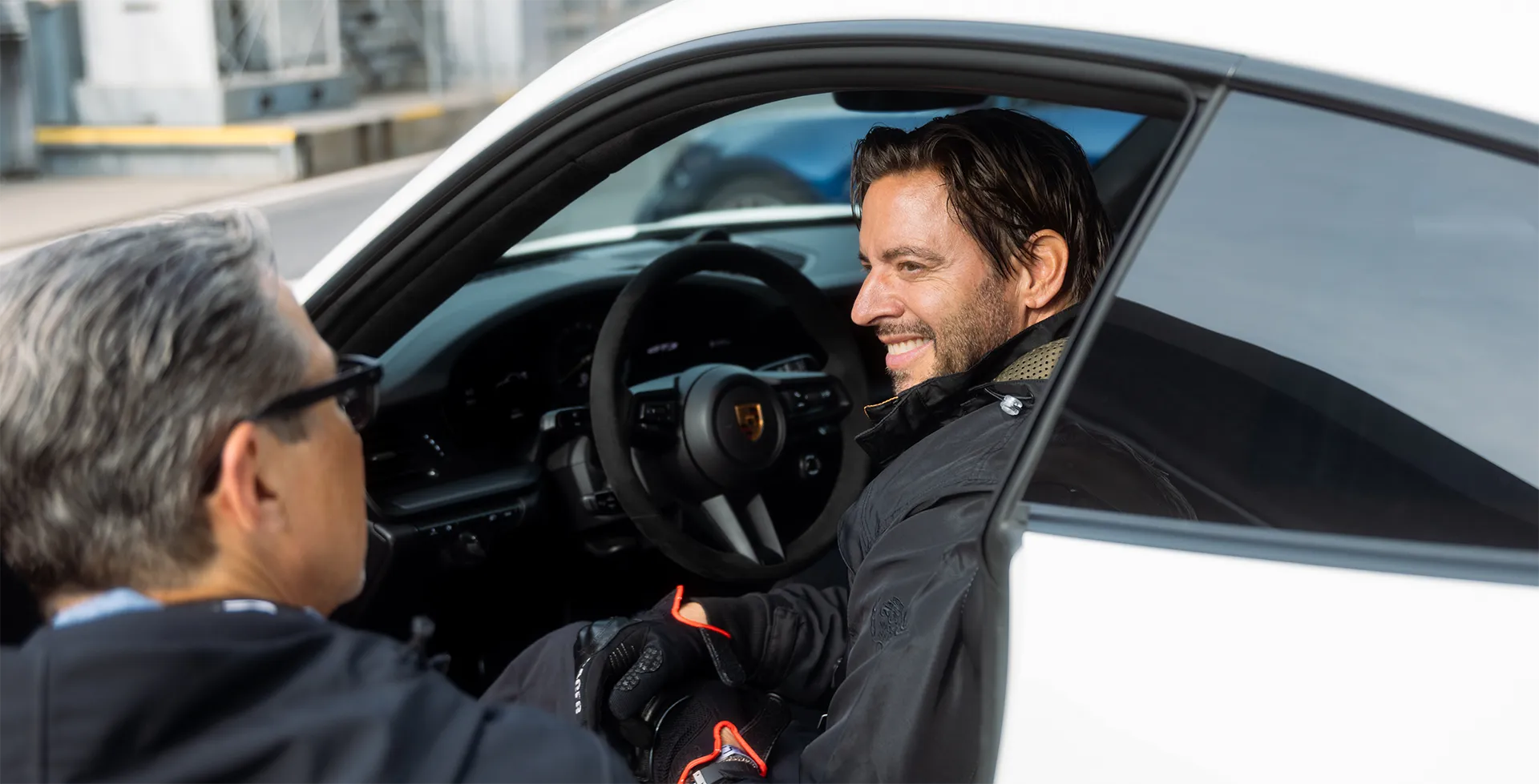 Mann mit schwarzen Handschuhen sitzt in einem weißen Porsche und lächelt während er mit einem anderen Mann spricht.