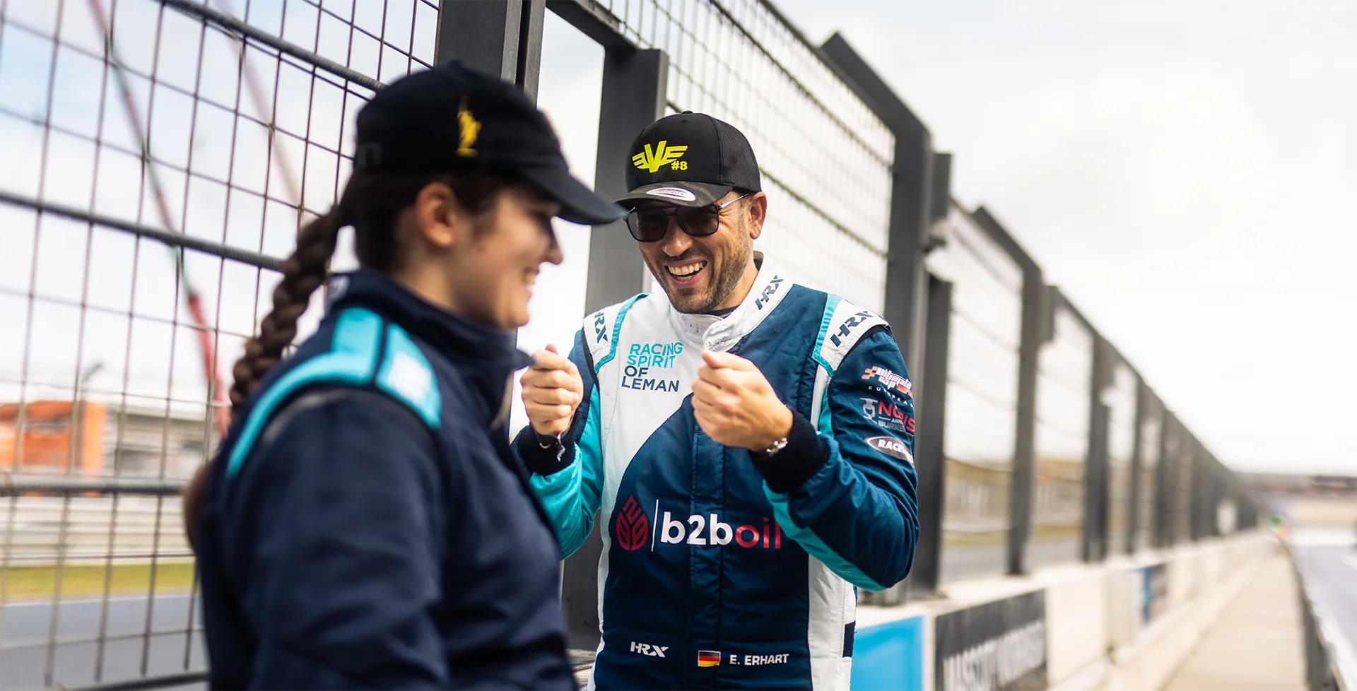 Rennfahrer in dunkelblauem Overall und schwarzer Mütze lacht und hält die Fäuste in Siegerpose neben einer Frau in dunkler Kleidung vor einem Zaun am Rennstreckenrand.