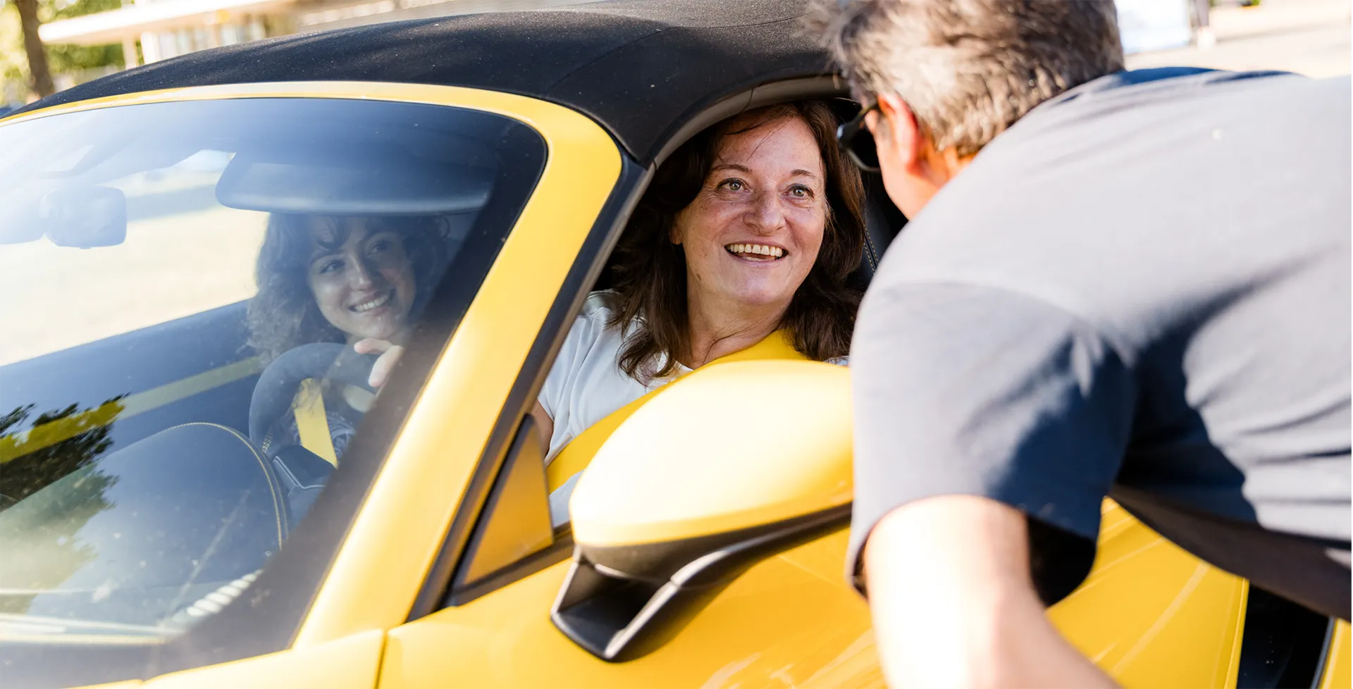 Eine lächelnde Frau sitzt in einem gelben Cabrio, während ein Mann sich zum Auto lehnt und mit ihr spricht.