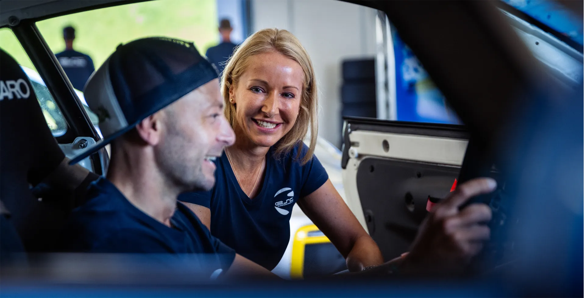 Frau lächelt und spricht mit einem Mann, der ein schwarzes Cap trägt, während sie in einem Auto sitzen.
