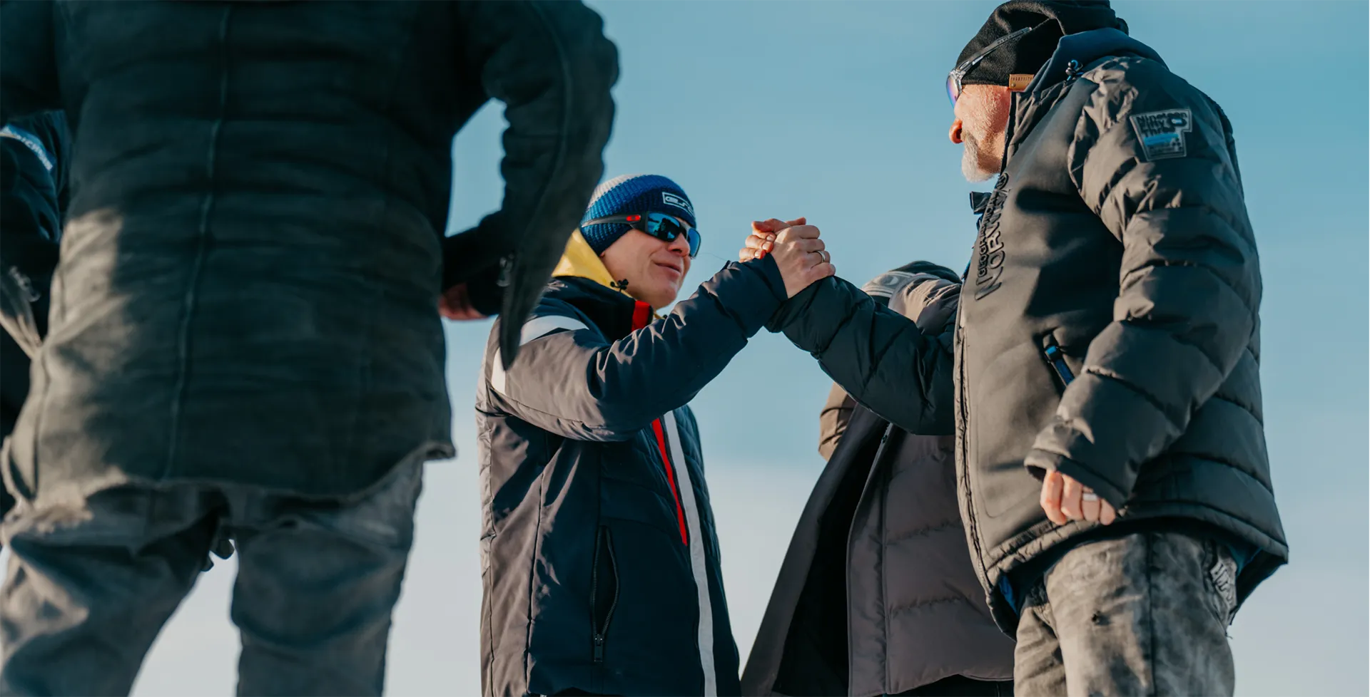 Zwei Männer in Winterjacken begrüßen sich mit einem Handschlag im Freien bei blauem Himmel.