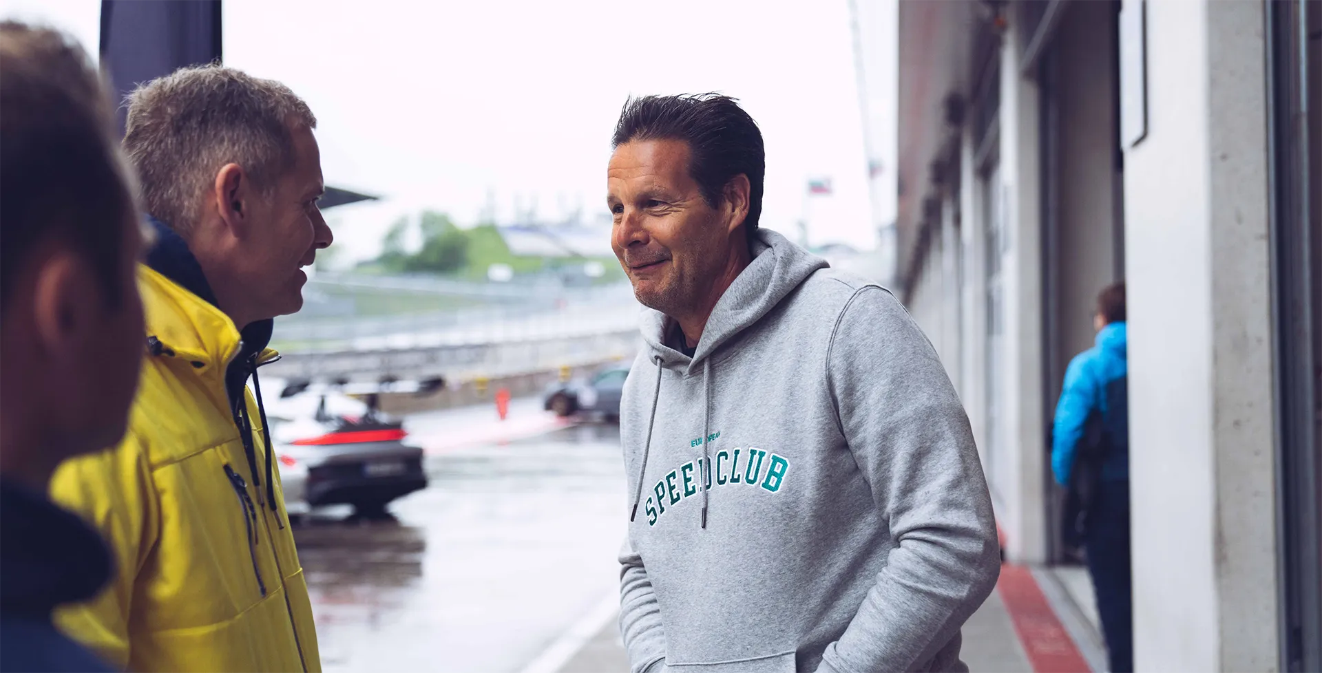 Zwei Männer stehen unter einem Überdach neben einer Rennstrecke, einer trägt eine gelbe Jacke, der andere einen grauen Hoodie mit der Aufschrift Speedclub.