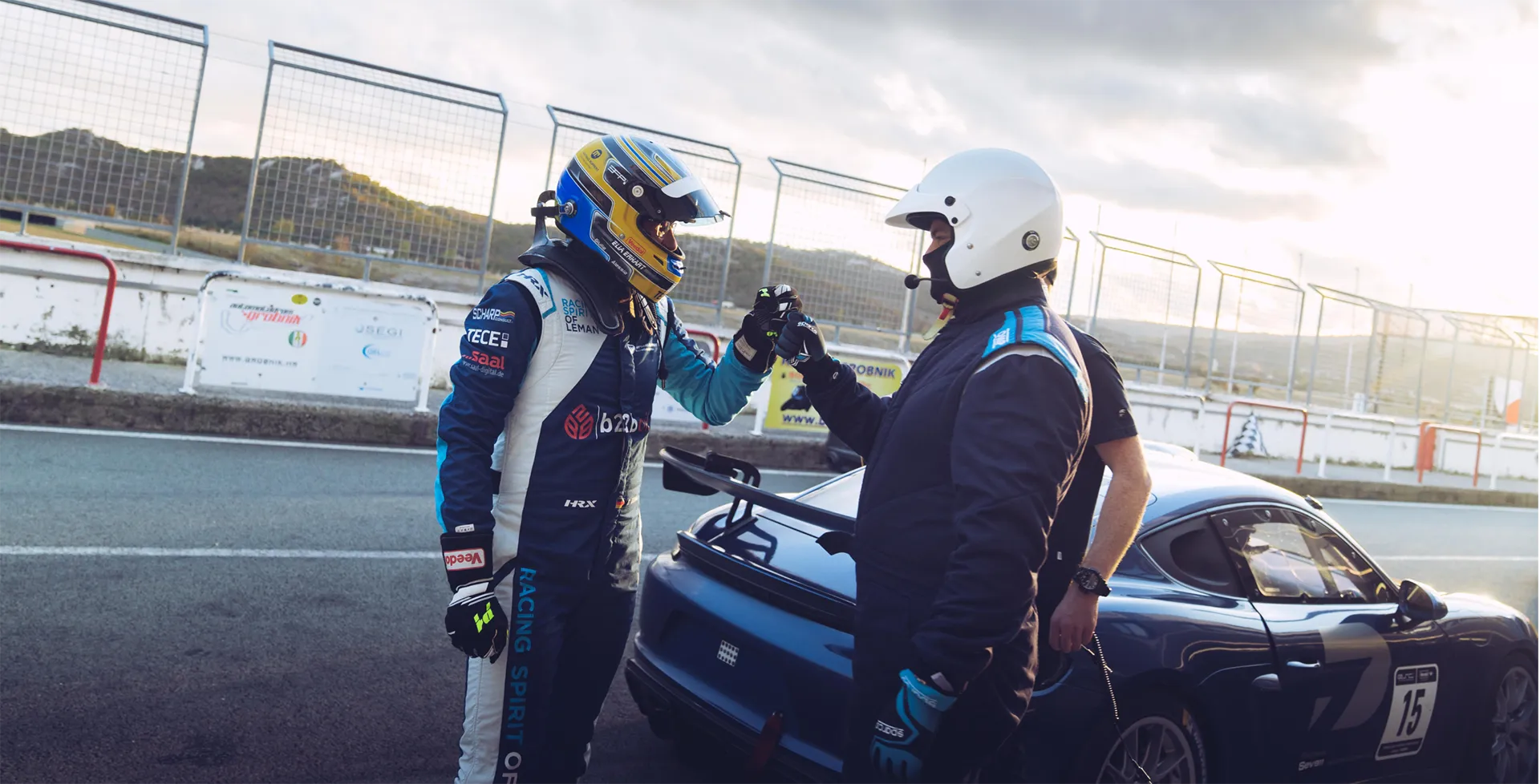 Zwei Rennfahrer in Schutzanzügen und Helmen geben sich auf einer Rennstrecke einen Handschlag neben einem blauen Rennwagen.