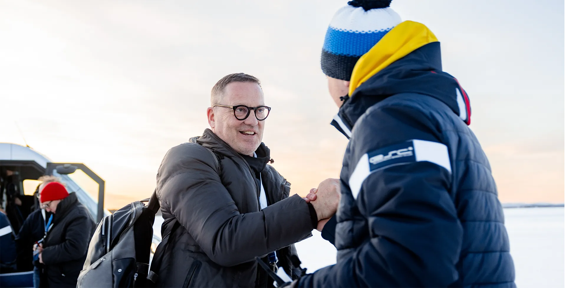 Zwei Männer in Winterjacken, die sich im Freien bei Sonnenuntergang herzlich die Hand schütteln.