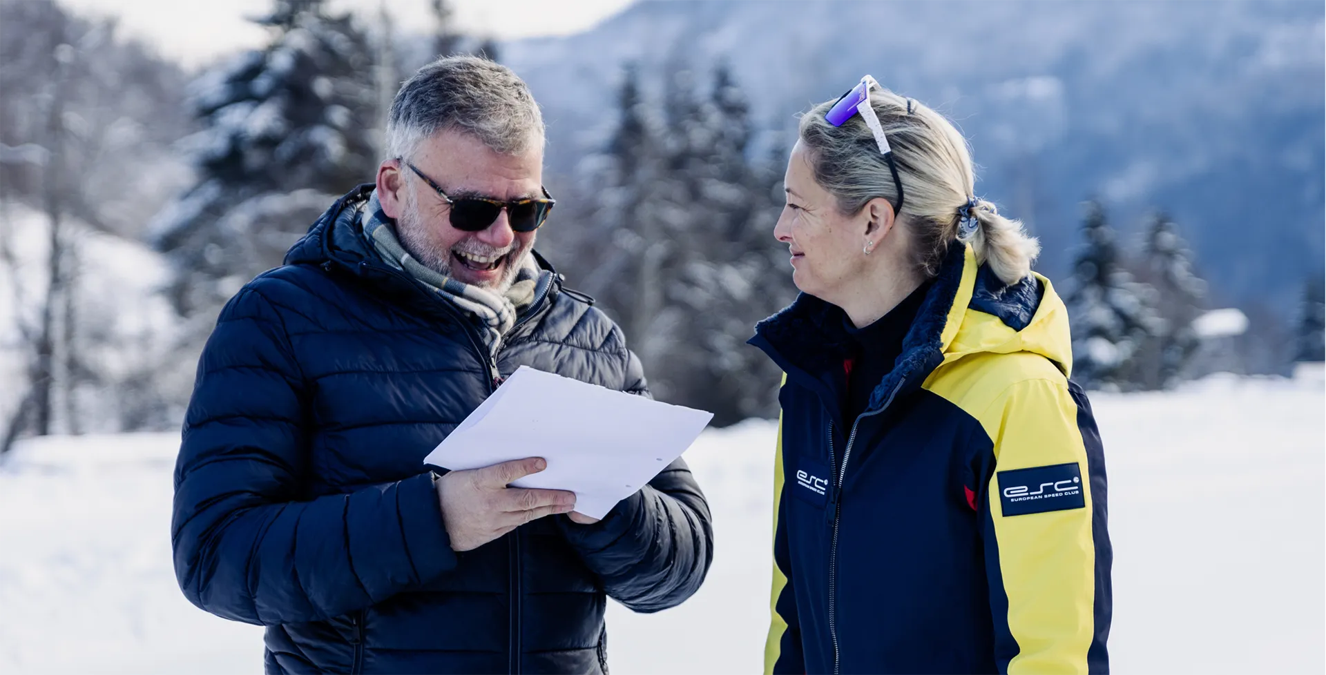 Ein lächelnder Mann in einer dunklen Winterjacke und eine Frau in einer blauen und gelben Jacke unterhalten sich draußen im Schnee.