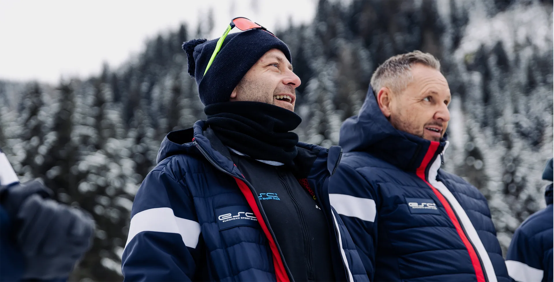 Zwei Männer in blauen ESC-Jacken stehen vor einem schneebedeckten Wald und blicken nach rechts.