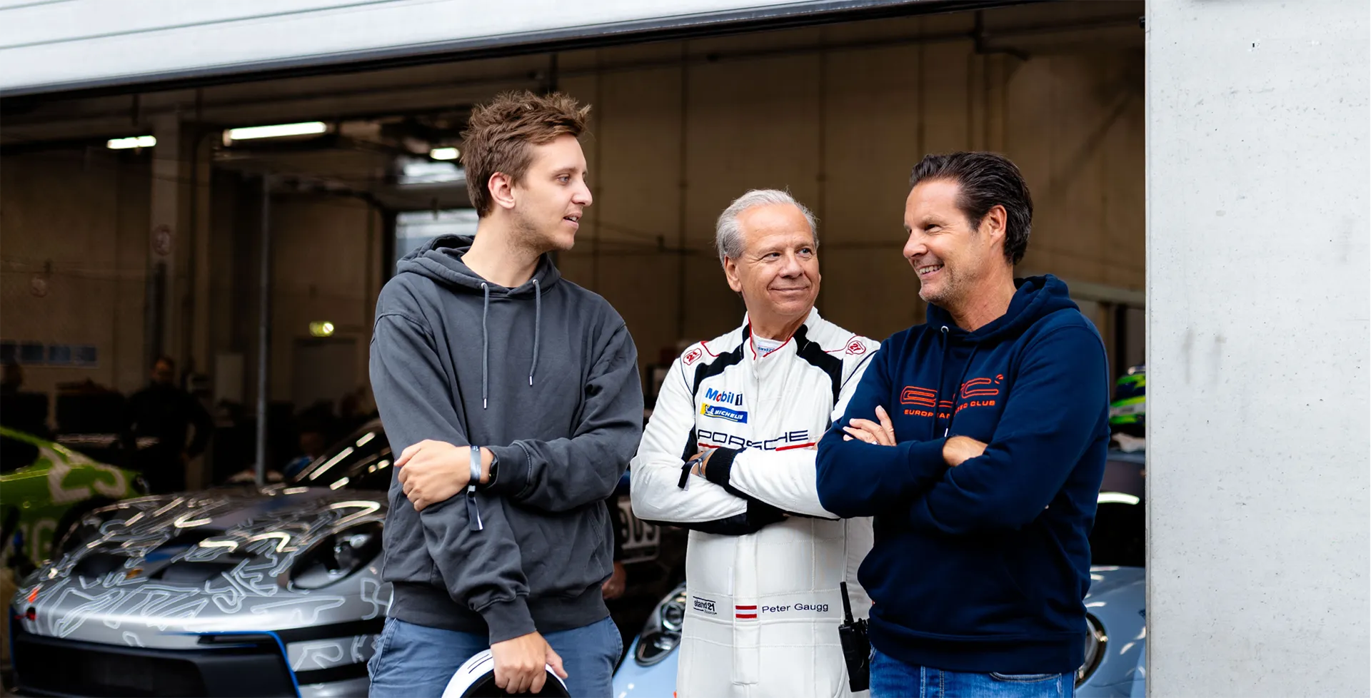Drei Männer stehen in einer Garage vor Rennwagen, einschließlich eines Mannes im Porsche-Rennanzug mit dem Namensschild Peter Gaugg.