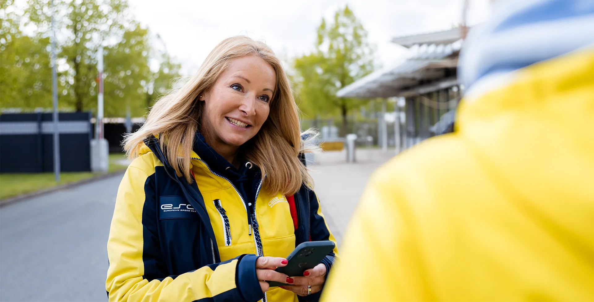 Frau in gelb-schwarzer Jacke hält ein Smartphone und lächelt, während sie im Freien mit einer anderen Person spricht.