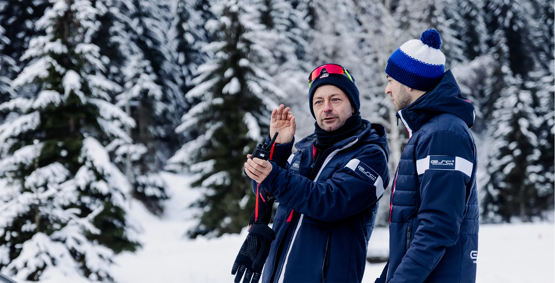 Zwei Männer in Winterjacken und Mützen stehen im verschneiten Wald, einer hält ein Funkgerät und erklärt etwas.