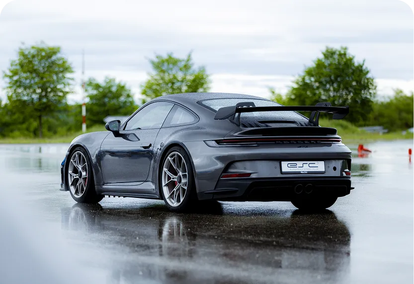 Grauer Porsche 911 GT3 mit großem Heckflügel auf nasser Straße vor grünem Hintergrund.