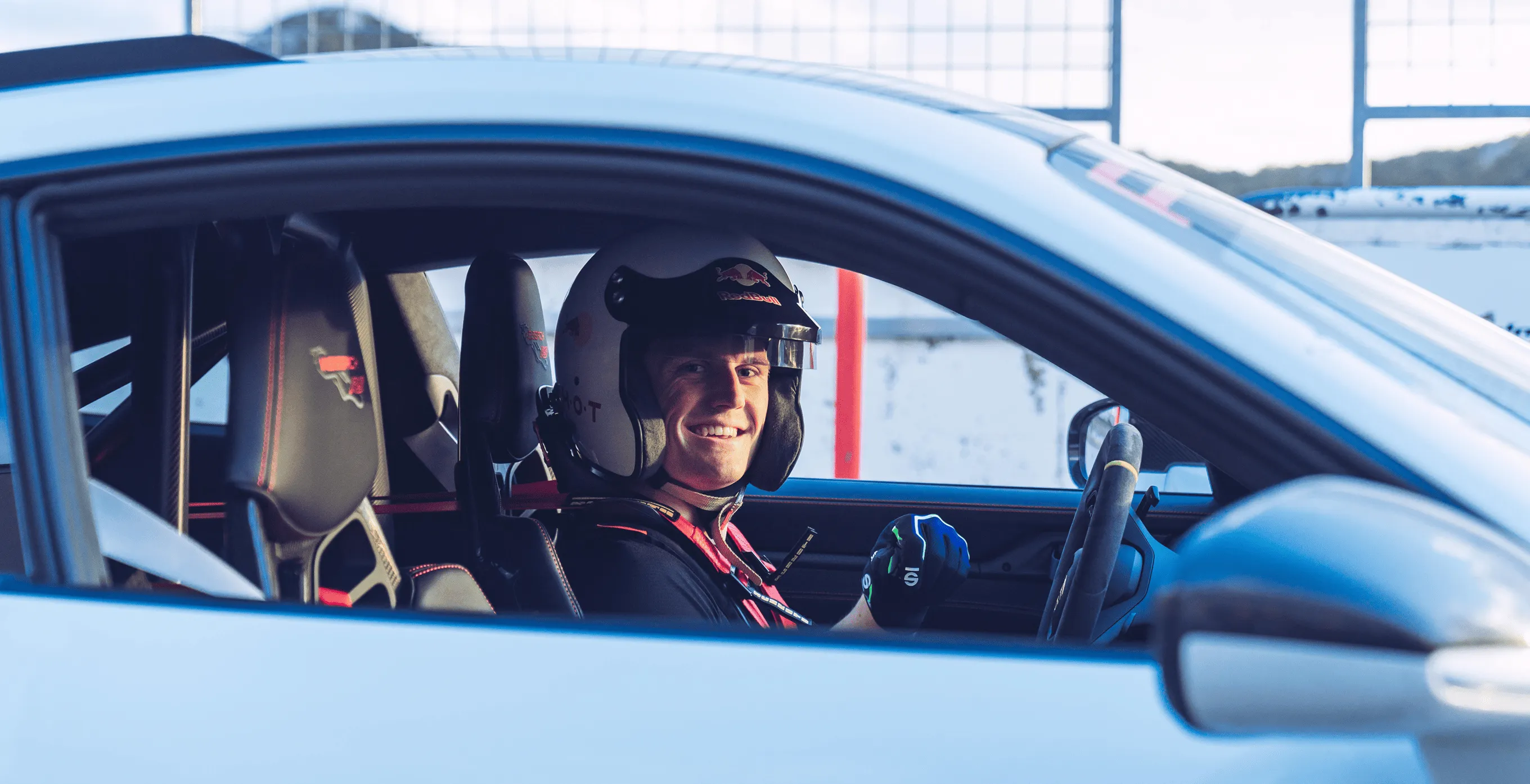Rennfahrer mit Helm und Handschuhen sitzt lächelnd im weißen Rennwagen.