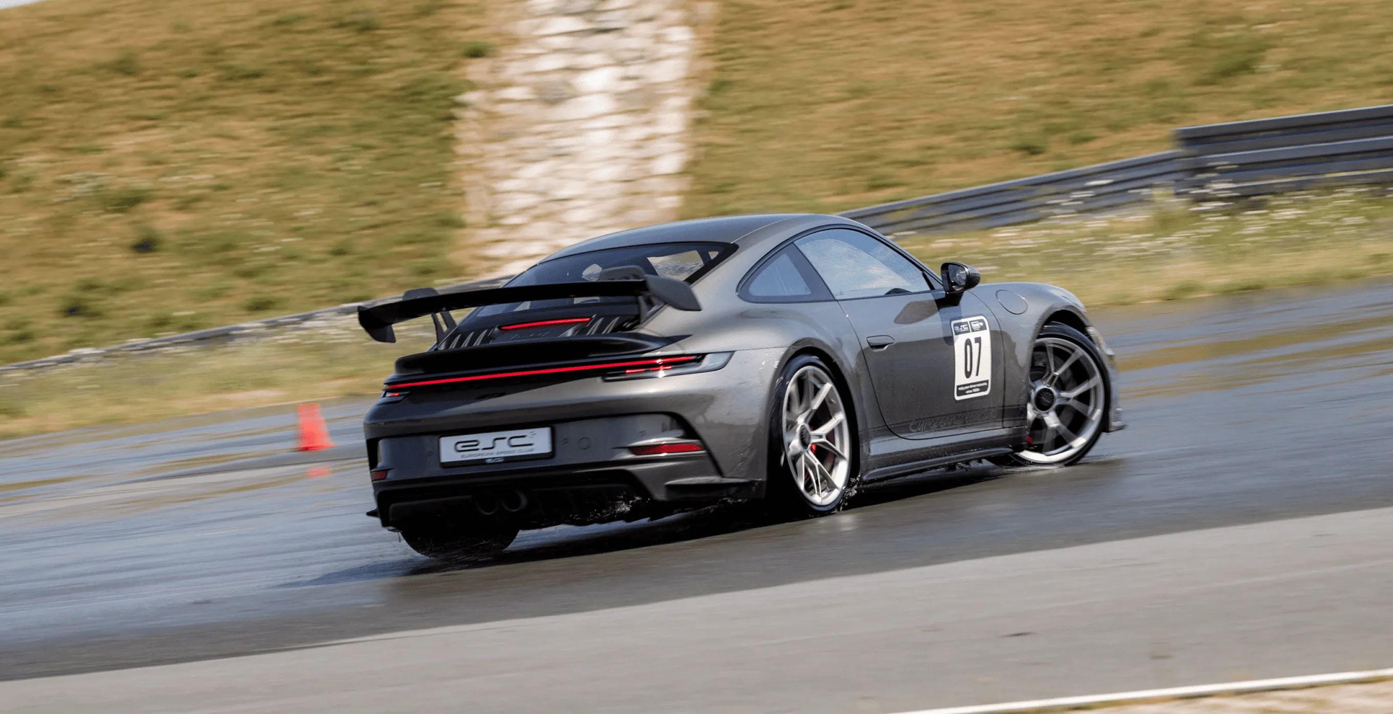 Dunkelgrauer Sportwagen mit Rennnummer 07 driftet auf nasser Rennstrecke mit orangefarbener Pylon im Hintergrund.