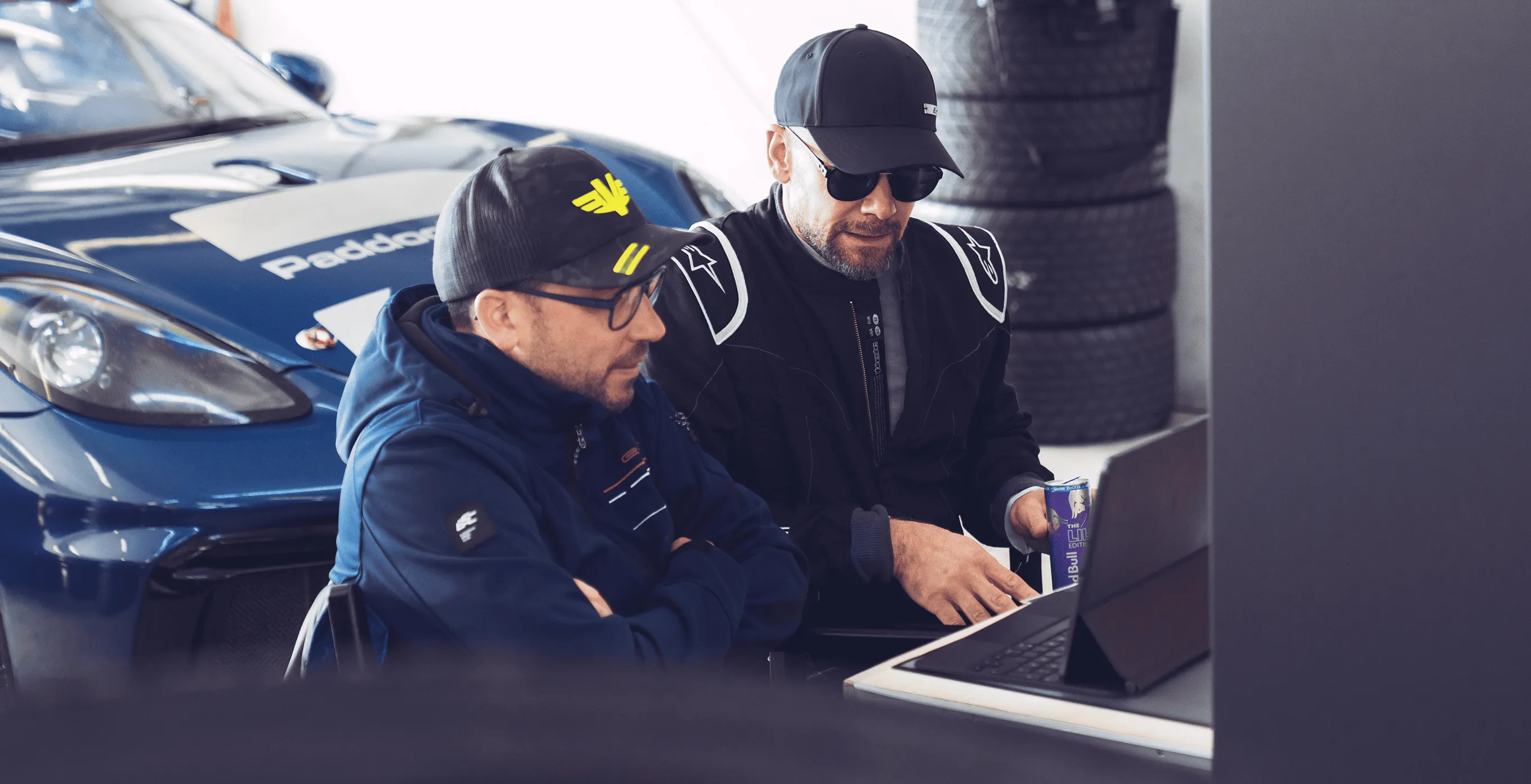 Zwei Männer in Motorsportkleidung sitzen vor einem Laptop, daneben ein blauer Rennwagen und gestapelte Reifen.