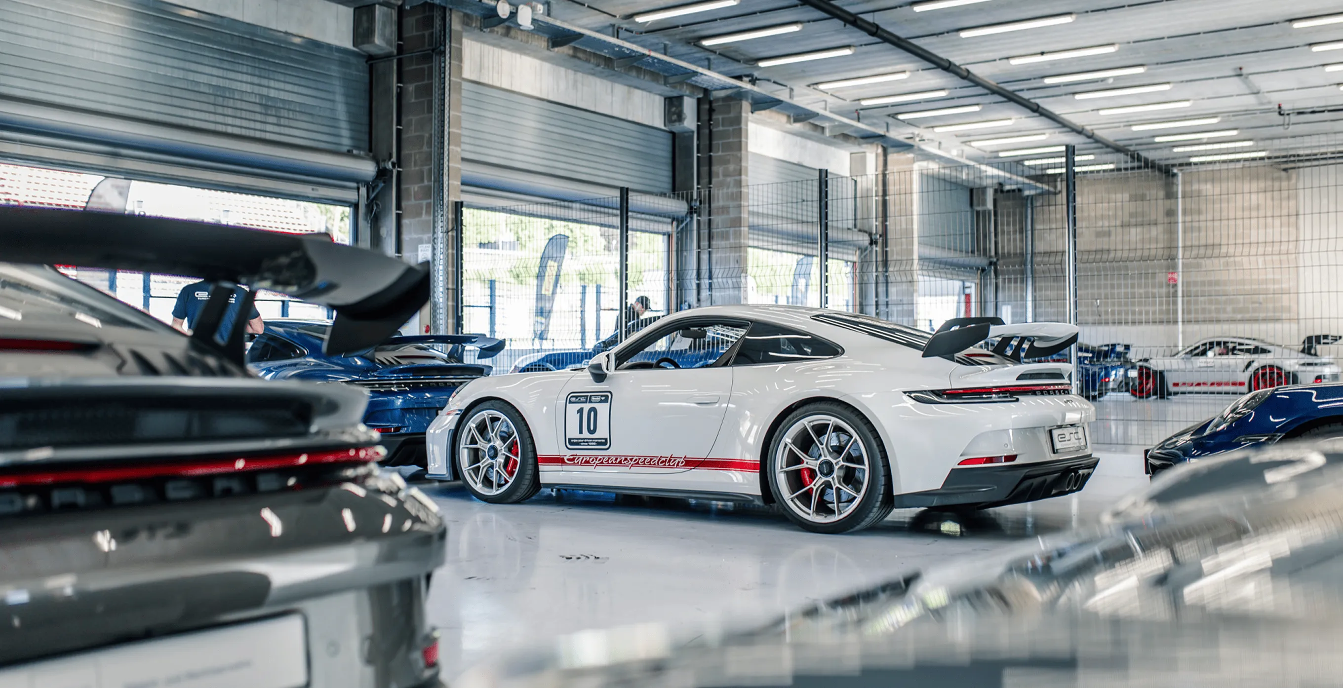 Weißer Sportwagen mit der Nummer 10 und rotem Streifen in einer Garage voller weiterer Sportwagen.