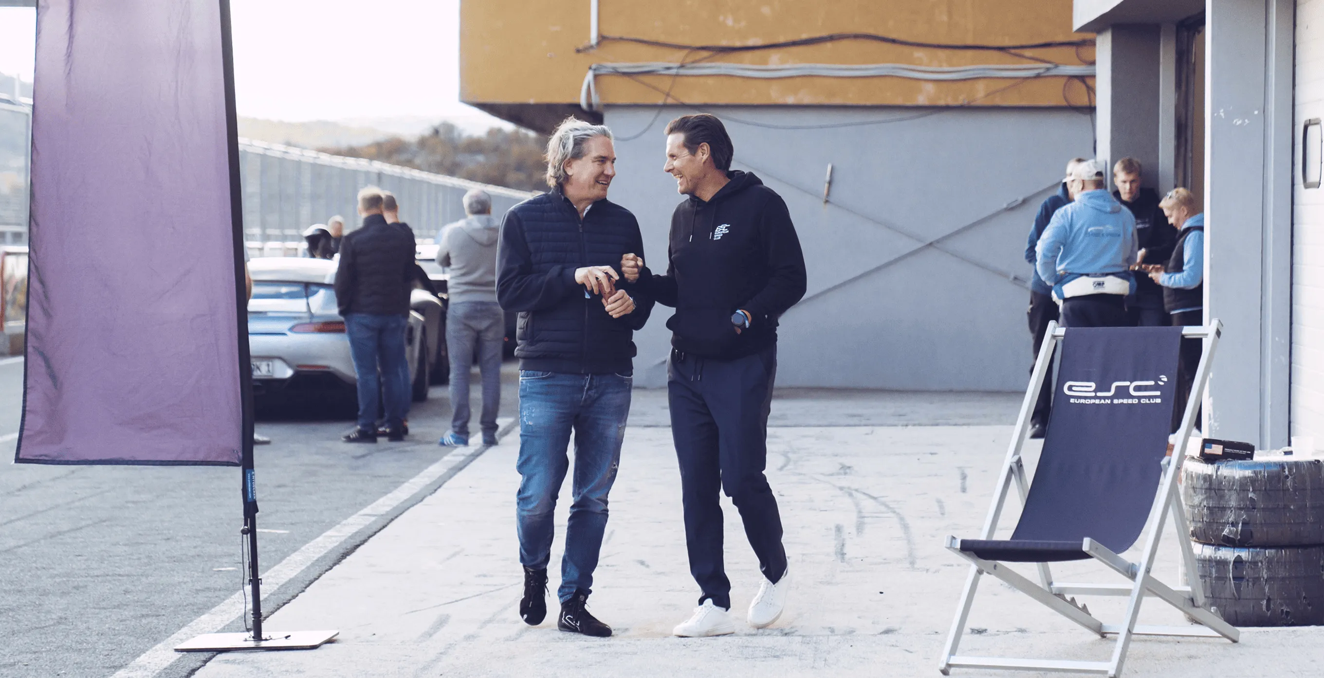 Zwei Männer in Freizeitkleidung lächeln und unterhalten sich auf einer Rennstrecke mit einem Porsche im Hintergrund und einer klappbaren Stuhlanzeige mit European Speed Club Schriftzug.