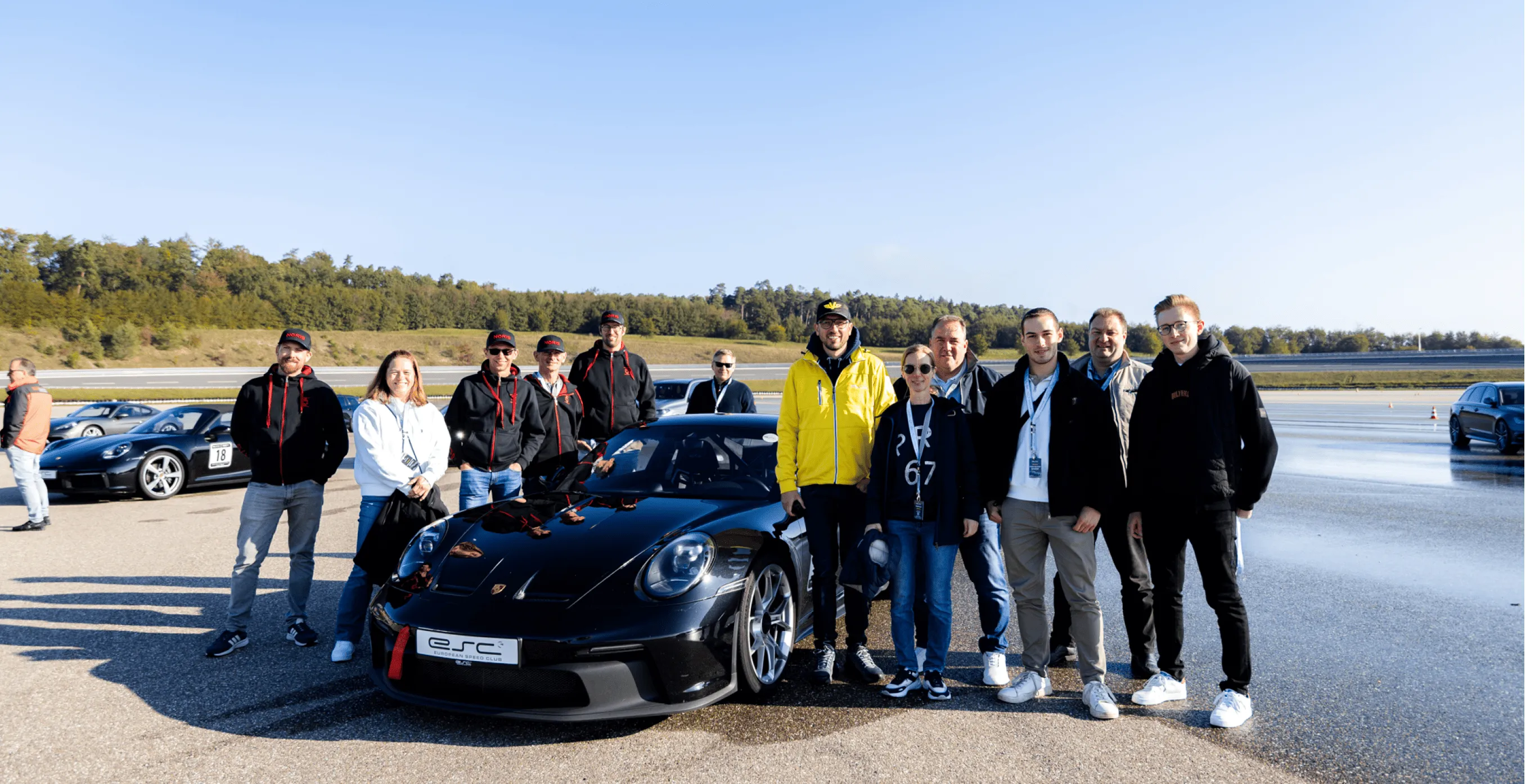 Gruppe von Menschen posiert bei sonnigem Wetter vor einem schwarzen Porsche 911 auf einem Rennstreckenparkplatz.