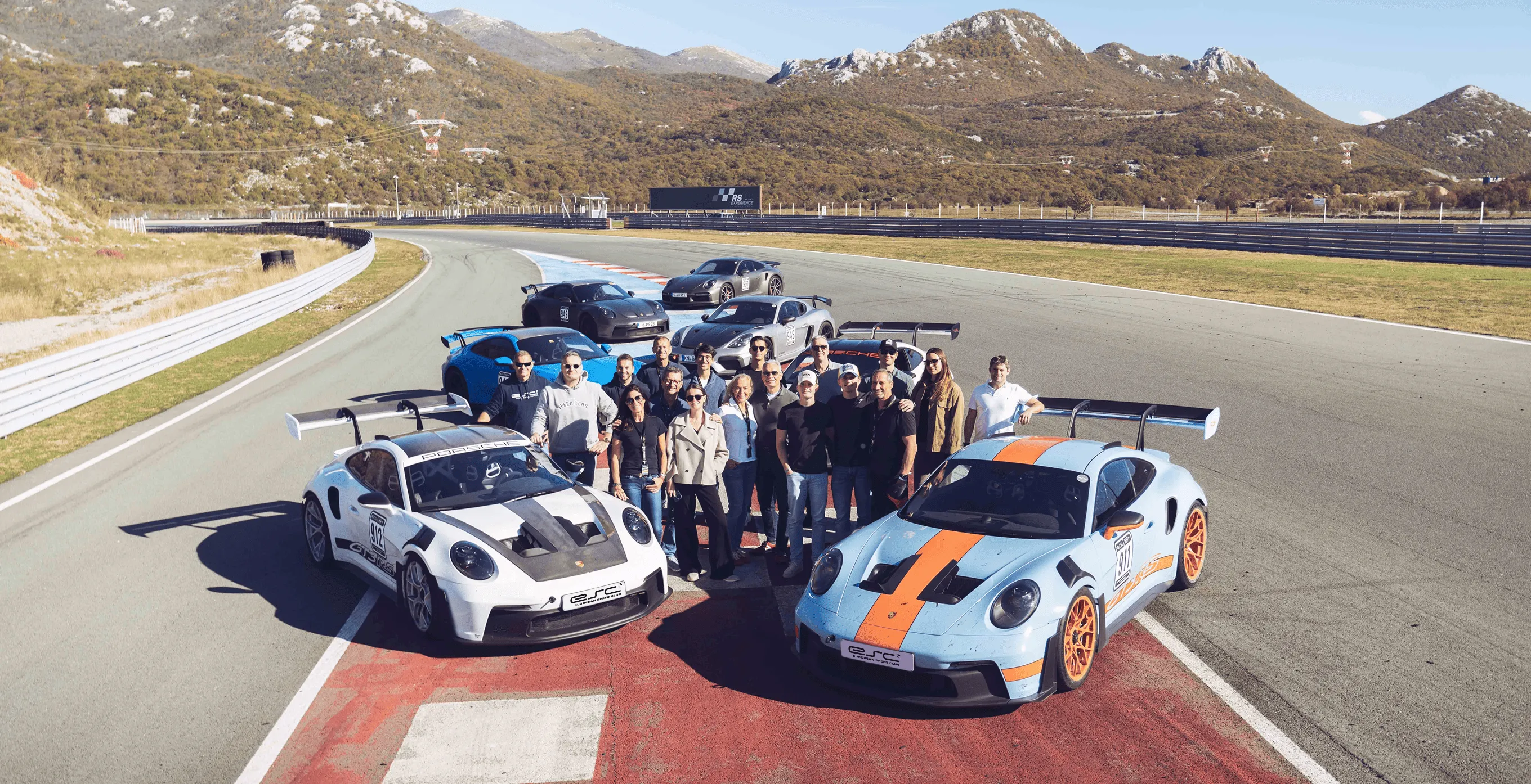 Gruppe von Menschen, die zwischen sechs Rennsport-Porsche auf einer Rennstrecke mit Bergen im Hintergrund posieren.
