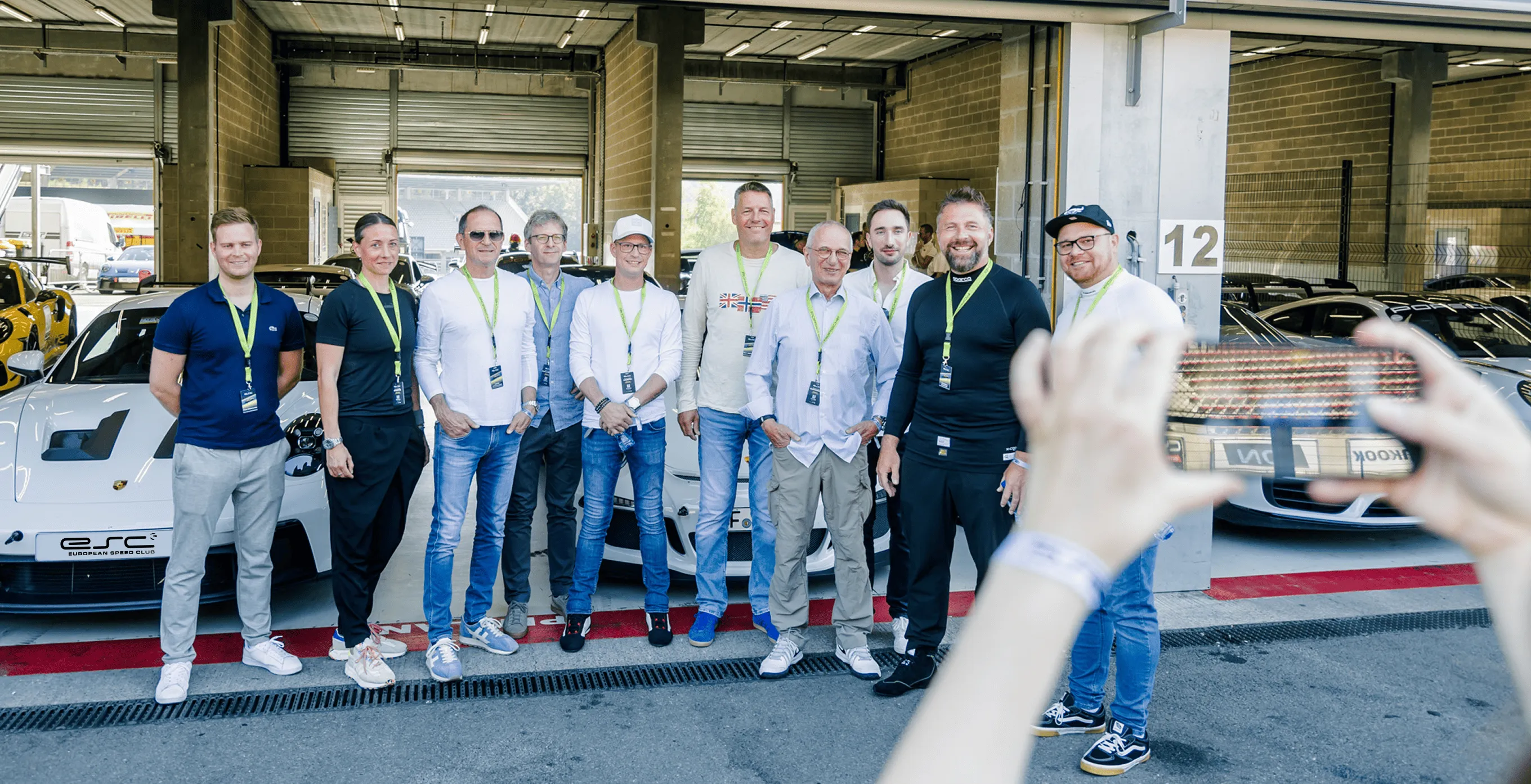 Zehn Menschen mit grünen Namensschildern posieren vor weißen Sportwagen in einer Garage, während eine Person im Vordergrund ein Foto mit einem Smartphone macht.