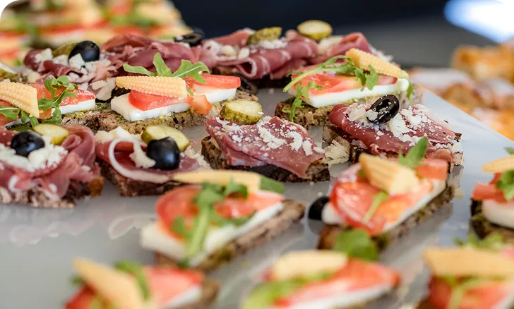 Verschiedene belegte Brotscheiben mit Tomaten, Schinken, Gurken, Mozzarella, schwarzer Olive und Babymais auf einer Platte.