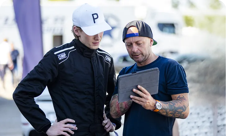 Zwei Männer blicken gemeinsam auf ein Tablet, einer trägt einen weißen Baseballcap und ein schwarzes Rennfahrer-Outfit, der andere eine rückwärts getragene Kappe und ein T-Shirt mit Tattoos an den Armen.