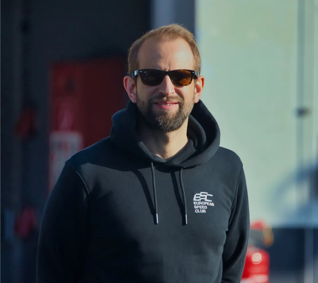 Mann mit Sonnenbrille und schwarzem Kapuzenpullover mit Aufschrift 'European Speed Club' steht draußen bei Sonnenlicht.