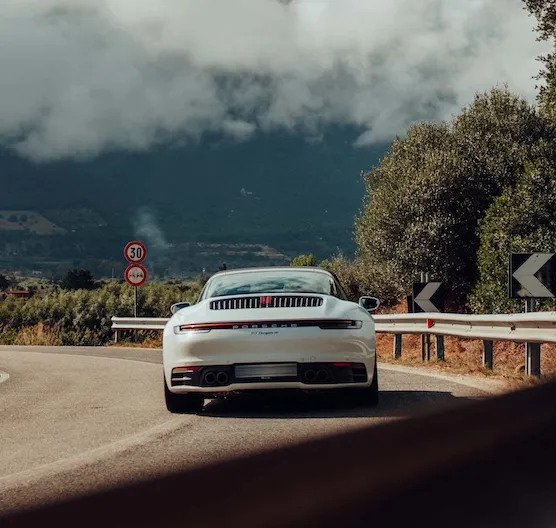 Weiße Porsche Sportwagenschwärze fährt auf einer kurvigen Bergstraße mit Schildern und Bewuchs bei bewölktem Himmel.