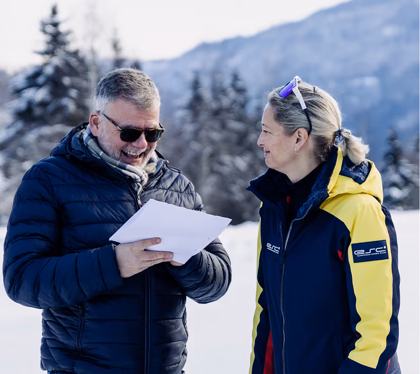 Two people in winter clothes are talking outside in front of snow-covered trees and mountains.