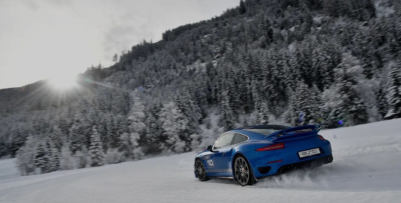 Blauer Sportwagen fährt auf schneebedeckter Straße mit verschneiten Bäumen und Berg im Hintergrund.